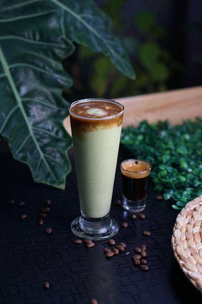 Exquisite blend of matcha and espresso in a tall glass with coffee beans and green leaves accenting the scene.