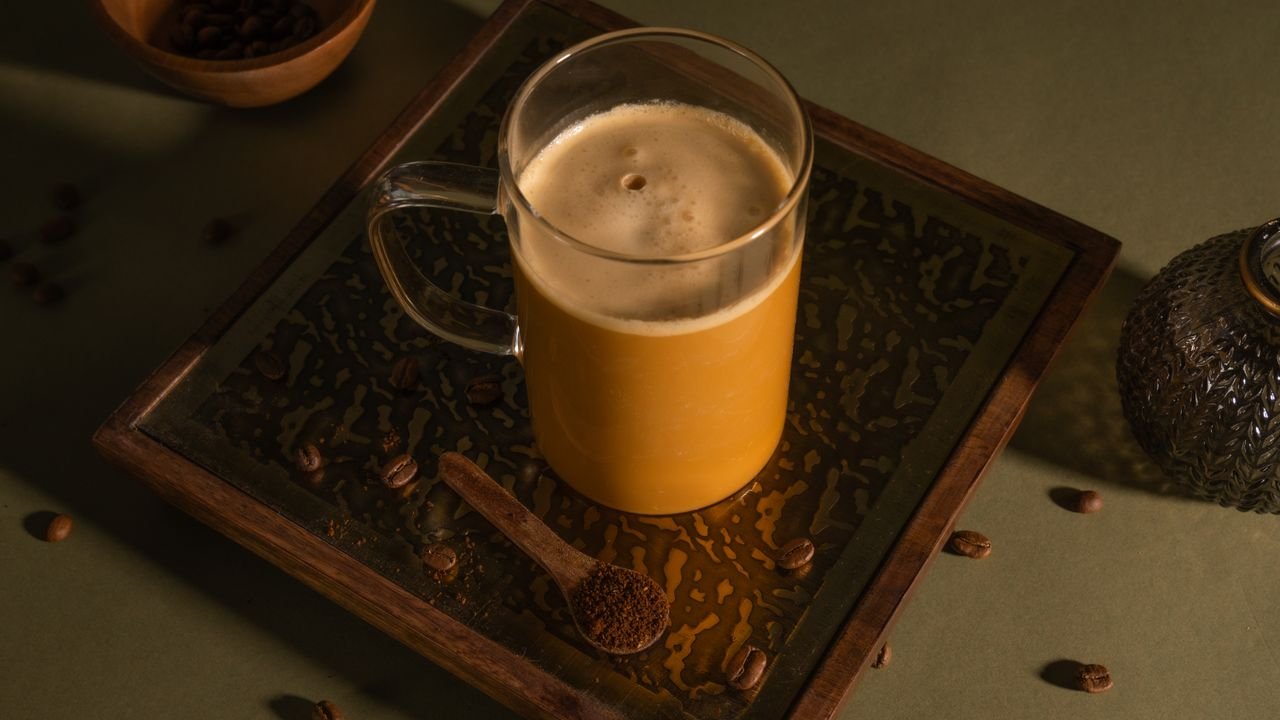 A glass cup filled with coffee on a decorative tray beside a spoon of ground coffee and scattered beans.